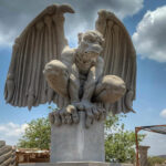 A large, light-colored cantera stone sculpture of a crouching gargoyle with outstretched wings, set against a blue sky with some clouds.