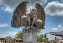 A large, light-colored cantera stone sculpture of a crouching gargoyle with outstretched wings, set against a blue sky with some clouds.
