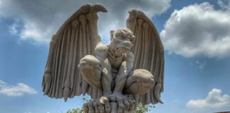 A large, light-colored cantera stone sculpture of a crouching gargoyle with outstretched wings, set against a blue sky with some clouds.