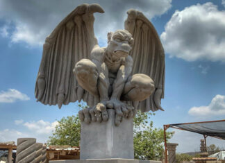 A large, light-colored cantera stone sculpture of a crouching gargoyle with outstretched wings, set against a blue sky with some clouds.