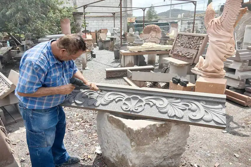 A skilled stonemason artisan demonstrating traditional hand-carving techniques on an ornate cantera stone frieze. Around him are other stone pieces in the an outdoor workshop