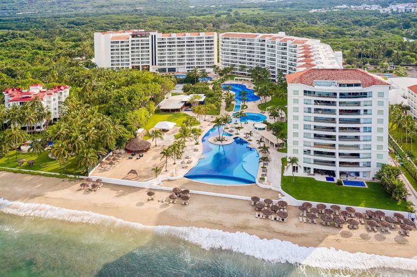 Aerial view of a large beachfront resort with multiple white buildings, winding pools, palm trees and straw-roofed beach cabanas.