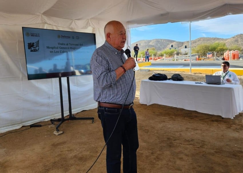Governor of Baja California Sur, Mexico Victor Castro standing under a large outdoor event tent with a microphone, in front of an oversized TV monitor displaying a slide presentation.
