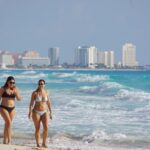 Two women in bikinis walk down the beach in Cancun, Quintana Roo, with the city skyline in the background