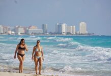 Two women in bikinis walk down the beach in Cancun, Quintana Roo, with the city skyline in the background