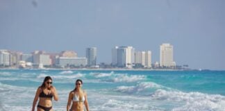 Two women in bikinis walk down the beach in Cancun, Quintana Roo, with the city skyline in the background