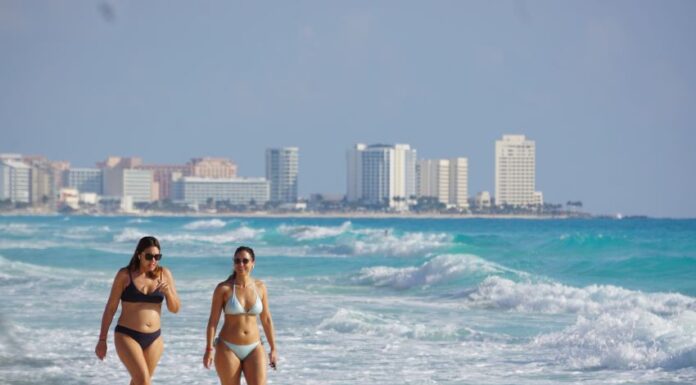 Two women in bikinis walk down the beach in Cancun, Quintana Roo, with the city skyline in the background