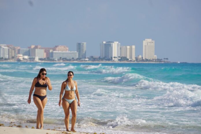Two women in bikinis walk down the beach in Cancun, Quintana Roo, with the city skyline in the background