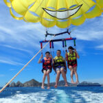 Three people strapped into a yellow parasail connected to a boat off camera. They are in the air slightly above the ocean.