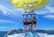 Three people strapped into a yellow parasail connected to a boat off camera. They are in the air slightly above the ocean.