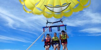 Three people strapped into a yellow parasail connected to a boat off camera. They are in the air slightly above the ocean.