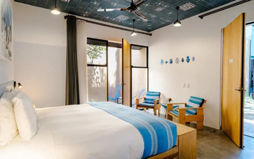 A brightly lit hotel bedroom featuring a large bed with white linens and a blue striped throw. The room has white walls and a striking dark blue ceiling with light-colored patterns. Two wooden chairs with blue and white striped cushions sit near an open door leading to an outdoor area with a blue hammock. Another open wooden door on the right reveals more of the exterior.