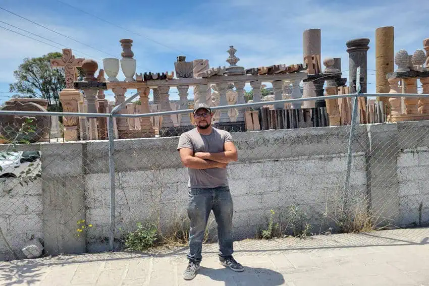 A Mexican man in his 20s or 30s stands outdoors with his arms crossed in front of his family's cantera artisan workshop, surrounded by diverse examples of their stone craftsmanship, including columns and balustrades.