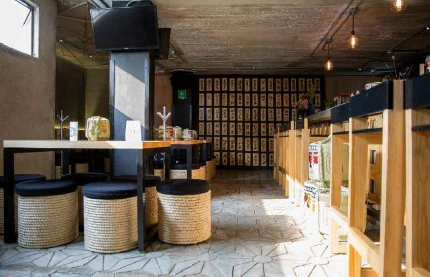 A bar with Japanese style light wooden decor and a wall in back with tiles decorated with Kanji symbols