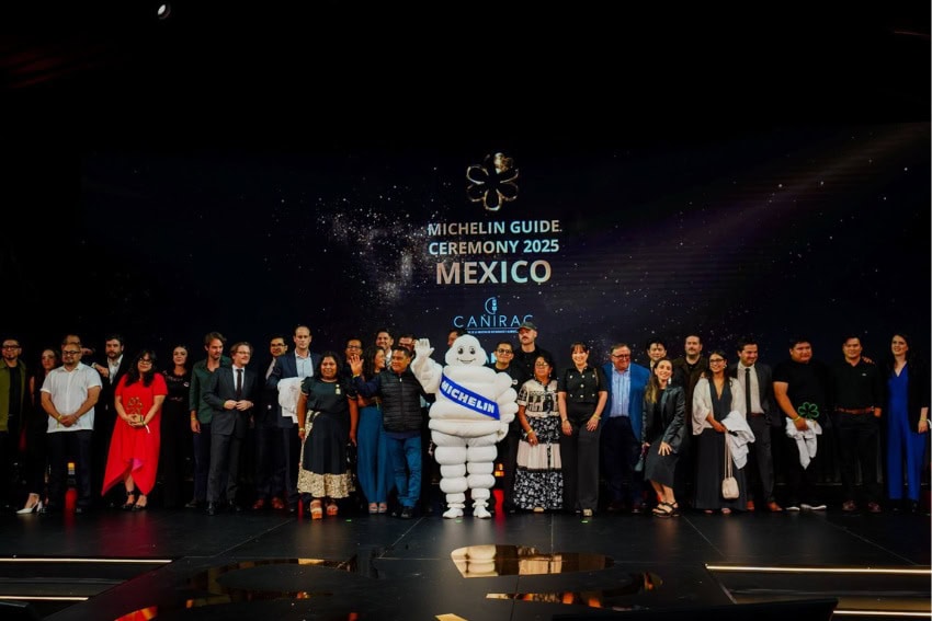 A group of about 30-40 people standing on a stage posing for a photo with a person in a Michelin Man costume at the center. Behind them is a legend on the stage's back wall that says Michelin Guide Ceremony 2025 Mexico