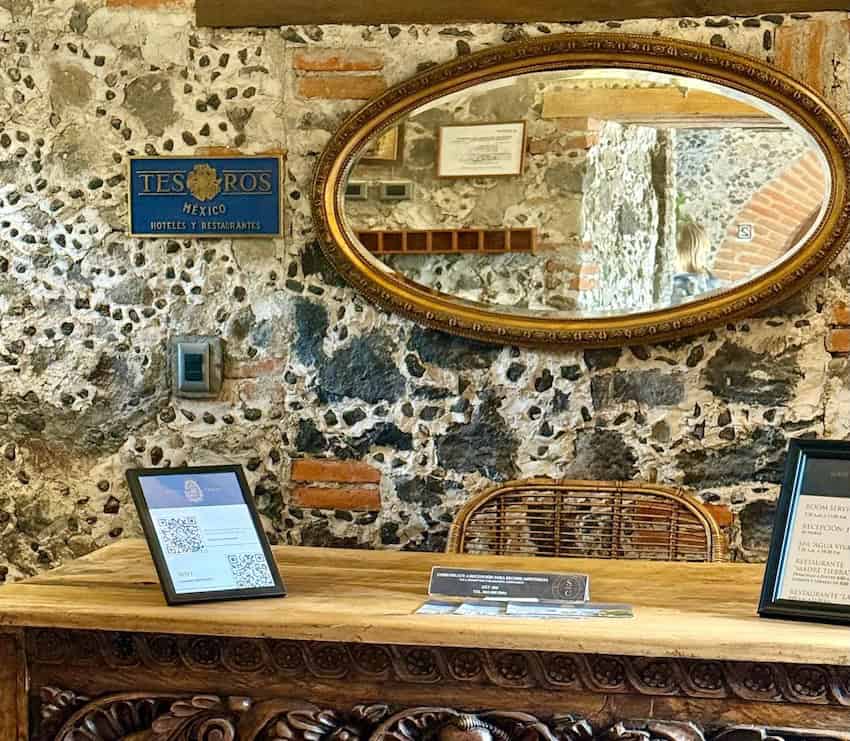 A room with stone and plaster walls in a historic hacienda in Mexico. The photo features a rustic antique wooden desk with baroque carvings. On the wall is an oval mirror with a gold leaf frame and a an antique-looking sign saying "Tesoros Mexico Hotels and Restaurants" in Spanish.