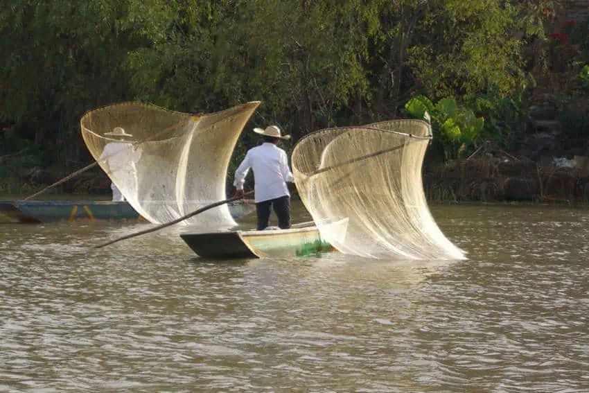 Two traditional fishermen in small boats on a calm lake, each holding up a large, distinctive "butterfly" fishing net made of light-colored netting stretched over wooden poles. The fisherman in the foreground is in a green and white boat, facing left, while the second fisherman is further back in a blue boat, mostly obscured. Lush green trees and foliage line the distant bank under a bright sky.