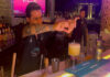 A bartender in a black bar apron focuses on squeezing a lime wedge over a yellow cocktail in a glass on the bar while another bartender looks on in the background.