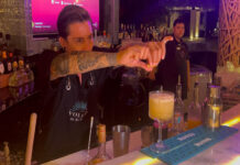 A bartender in a black bar apron focuses on squeezing a lime wedge over a yellow cocktail in a glass on the bar while another bartender looks on in the background.