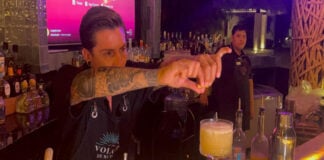 A bartender in a black bar apron focuses on squeezing a lime wedge over a yellow cocktail in a glass on the bar while another bartender looks on in the background.