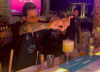 A bartender in a black bar apron focuses on squeezing a lime wedge over a yellow cocktail in a glass on the bar while another bartender looks on in the background.