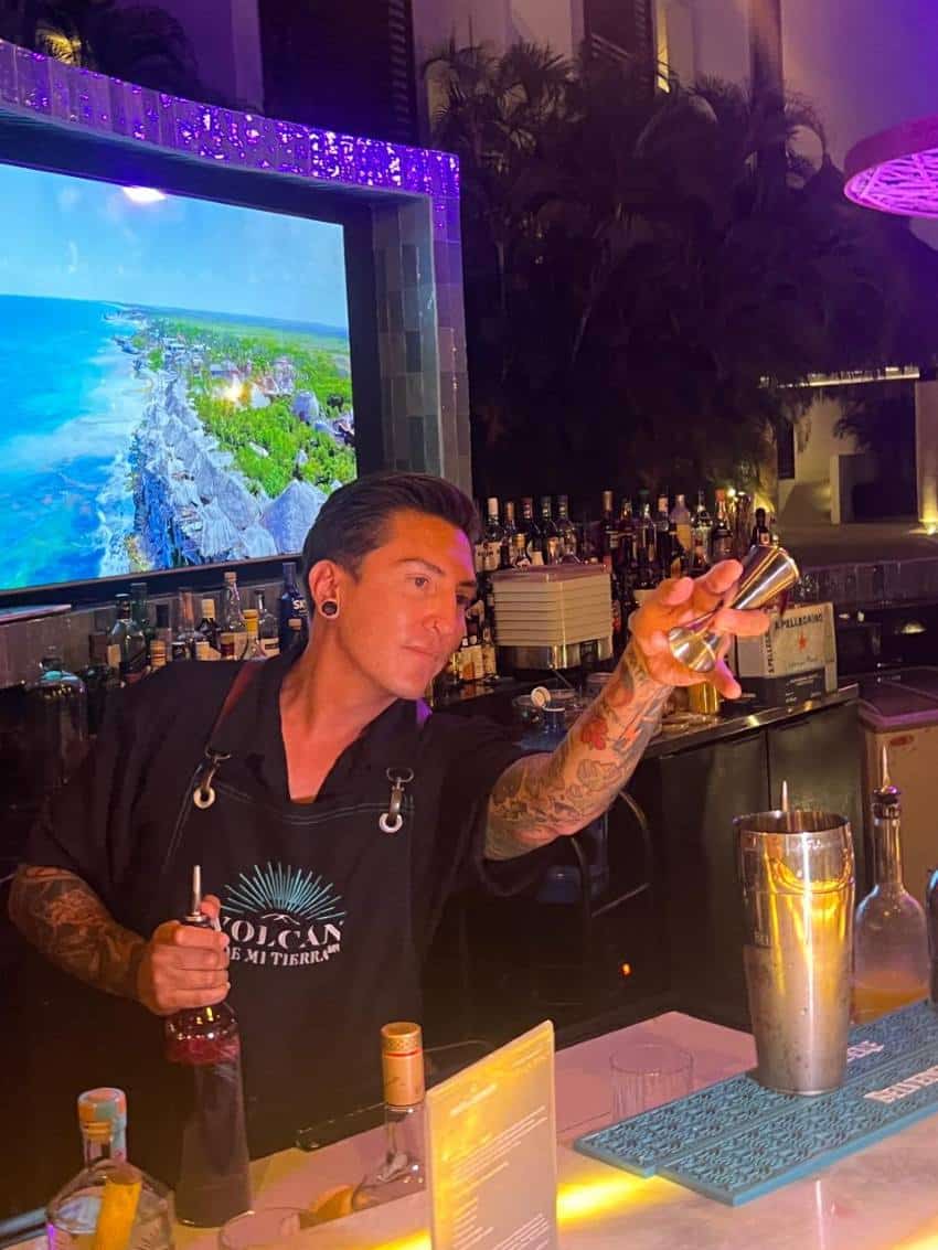 Bartender in a bar holding a bottle of liquor with a pouring spout on it and holding in the other hand a shot measure. He is gesturing with the shot measure to someone or something off camera as if taking an order from a patron.