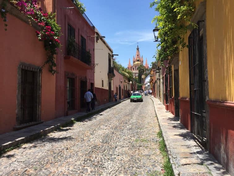 A taxi driving alone down a long, narrow downtown cobblestone street with multicolored adobe buildings with balconies and wooden doors