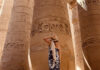 A tanned woman in a sundress and straw hat seen from the back as she looks up at massive pillars of an pre-Hispanic Indigenous structure in Mexico