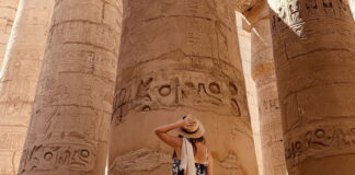 A tanned woman in a sundress and straw hat seen from the back as she looks up at massive pillars of an pre-Hispanic Indigenous structure in Mexico