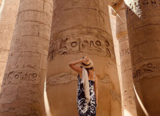 A tanned woman in a sundress and straw hat seen from the back as she looks up at massive pillars of an pre-Hispanic Indigenous structure in Mexico