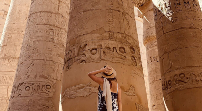 A tanned woman in a sundress and straw hat seen from the back as she looks up at massive pillars of an pre-Hispanic Indigenous structure in Mexico