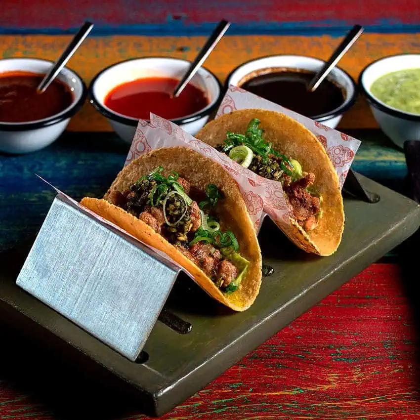 Tacos on a serving plate in front of four small dipping bowls of different salsas of different colors.