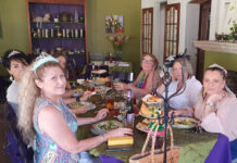 Six restaurant customers sitting at a long table decorated with satin tablecloths and featuring classic British teatime foods on traditional British tiered serving trays.