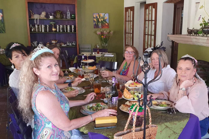 Six restaurant customers sitting at a long table decorated with satin tablecloths and featuring classic British teatime foods on traditional British tiered serving trays.