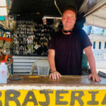 A man with a smiling expression and sunglasses pushed up on his head stands behind a counter at an outdoor key-making stand. Behind him, numerous keys are hanging, and the word "CERRAJERIA" (locksmith) is visible on the counter in yellow letters.