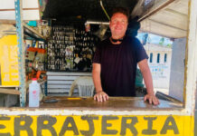 A man with a smiling expression and sunglasses pushed up on his head stands behind a counter at an outdoor key-making stand. Behind him, numerous keys are hanging, and the word "CERRAJERIA" (locksmith) is visible on the counter in yellow letters.