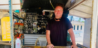 A man with a smiling expression and sunglasses pushed up on his head stands behind a counter at an outdoor key-making stand. Behind him, numerous keys are hanging, and the word "CERRAJERIA" (locksmith) is visible on the counter in yellow letters.