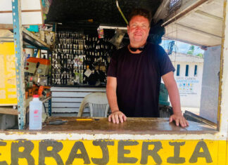 A man with a smiling expression and sunglasses pushed up on his head stands behind a counter at an outdoor key-making stand. Behind him, numerous keys are hanging, and the word "CERRAJERIA" (locksmith) is visible on the counter in yellow letters.