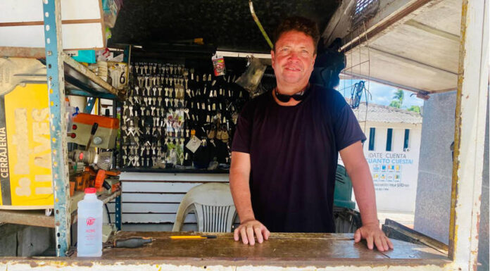 A man with a smiling expression and sunglasses pushed up on his head stands behind a counter at an outdoor key-making stand. Behind him, numerous keys are hanging, and the word "CERRAJERIA" (locksmith) is visible on the counter in yellow letters.