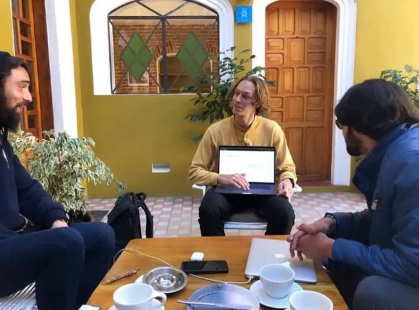 Three young men sitting at a coffee table in a Mexican courtyard. One has an open laptop in his lap and another has a laptop on the table next to his cup of coffee. The third is listening to the man with the laptop in his lap.