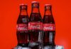 Three bottles of Coca-Cola on a pile of ice cubes and against a red background.