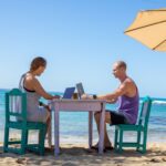 Man and woman work on laptops at a table on a beach