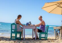Man and woman work on laptops at a table on a beach