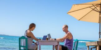Man and woman work on laptops at a table on a beach