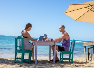Man and woman work on laptops at a table on a beach