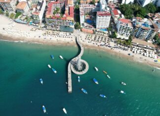 Aerial view of a winding pier with a circular end extending into clear blue-green ocean, with a sandy beach and a city skyline visible beyond.