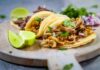 Mexican style tacos filled with meat, red onions and cilantro with some lime wedges on the side, all on a round, wooden charcuterie board.