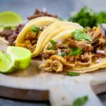 Mexican style tacos filled with meat, red onions and cilantro with some lime wedges on the side, all on a round, wooden charcuterie board.