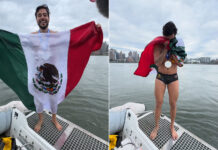 Mexican open water swimmer David Olvera stands on a boat in front of Manhattan Island in a swim suit, waving a Mexican flag