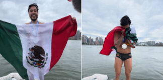 Mexican open water swimmer David Olvera stands on a boat in front of Manhattan Island in a swim suit, waving a Mexican flag